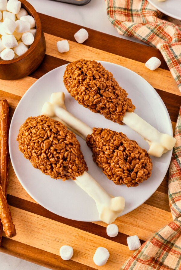 Cocoa krispie treats shaped around white chocolate dipped pretzel rods and marshmallow tips to look like turkey drumsticks. On a white plate, set on wooden cutting board, with marshmallows scattered around.