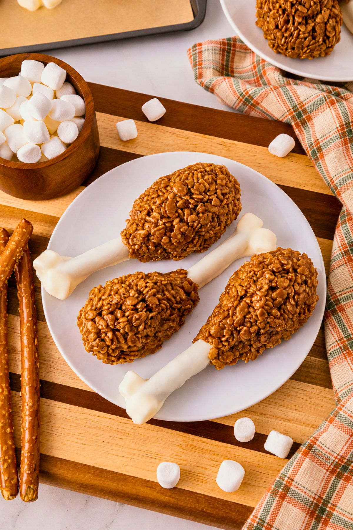 Cocoa krispie treats shaped around white chocolate dipped pretzel rods and marshmallow tips to look like turkey drumsticks. On a white plate, set on wooden cutting board, with marshmallows scattered around.