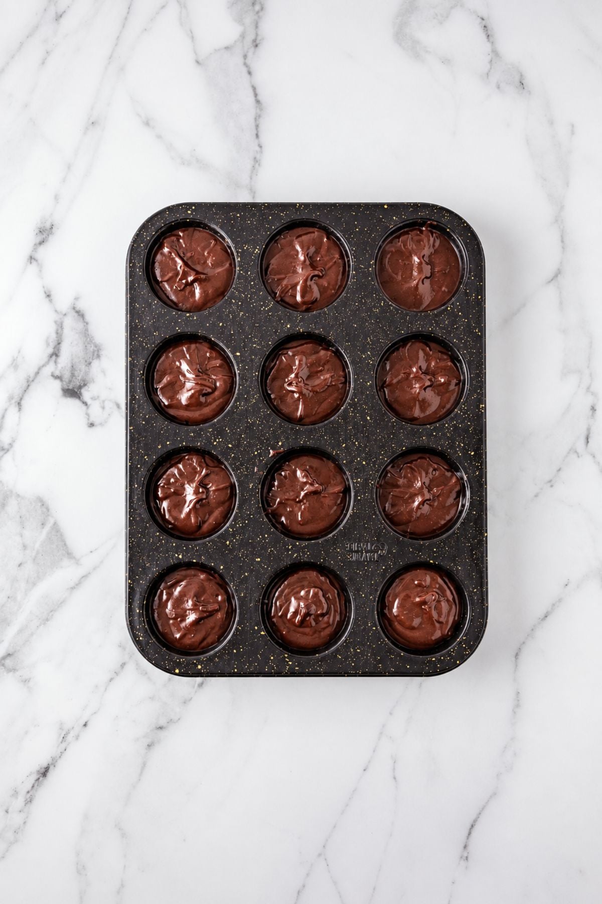 Muffin tin filled with brownie batter before baking, on a marble table.