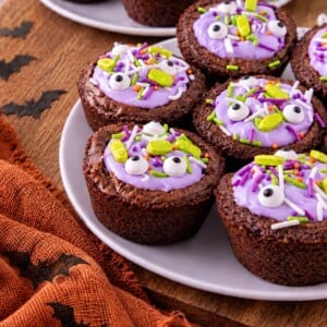 Brownies topped with purple frosting and Halloween sprinkles on a white plate with an orange linen on a wooden board.