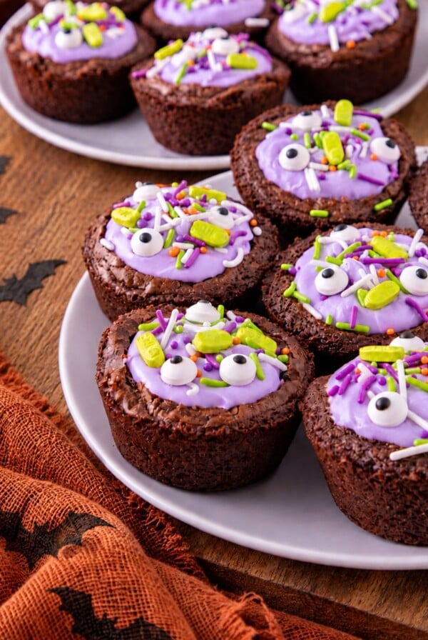 Brownies topped with purple frosting and Halloween sprinkles on a white plate with an orange linen on a wooden board.