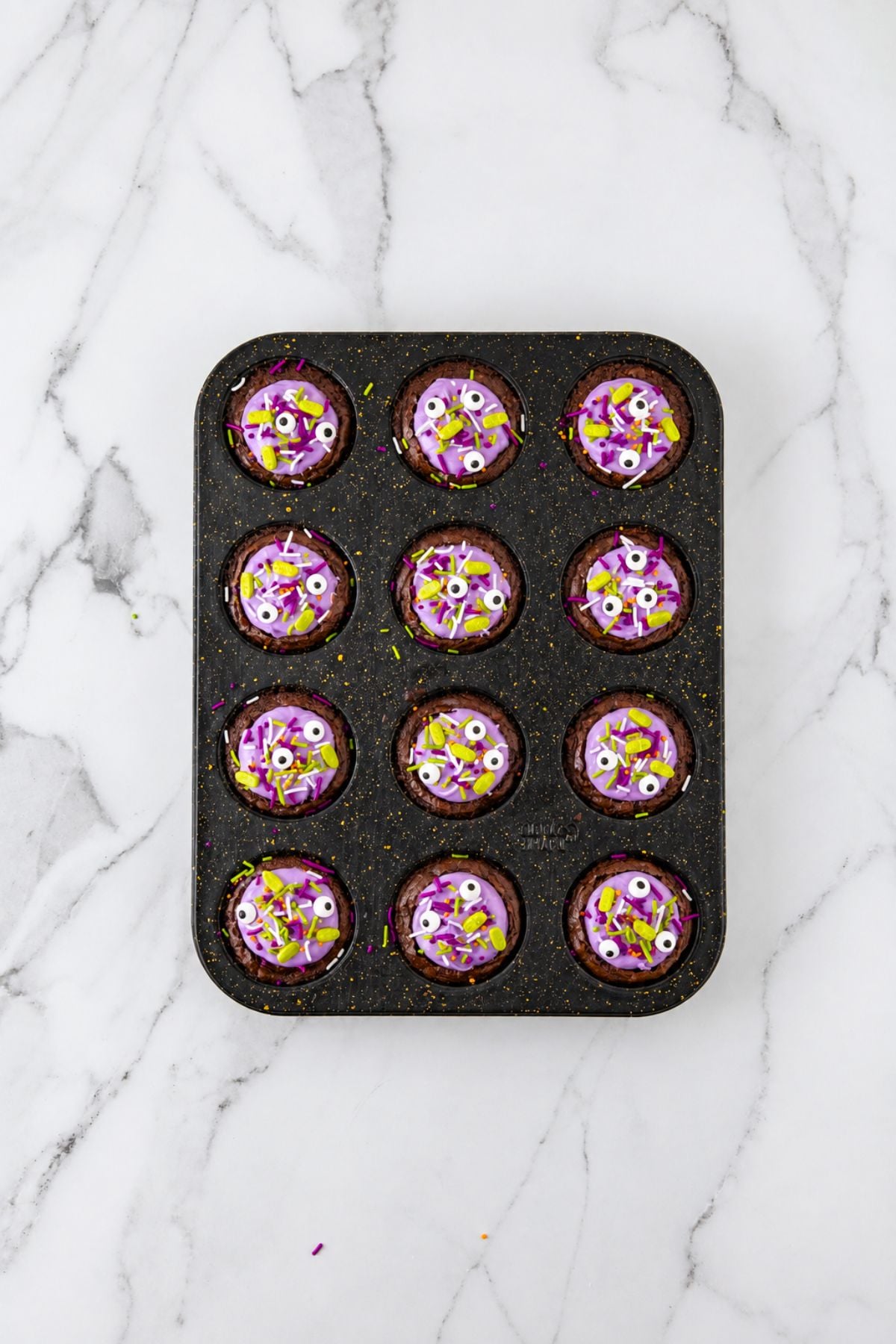 Brownie bites topped with purple frosting and sprinkles, in a muffin tin, on a marble table.