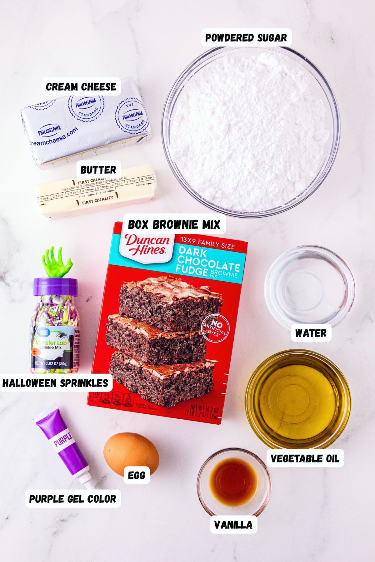 Ingredients of brownie mix, egg, cream cheese, powdered sugar, butter, coloring, vanilla, water, oil, and sprinkles, measured and labeled on a marble table.