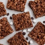 Chocolate rice krispie squares on a marble table with chips and mini marshmallows scattered on the table.