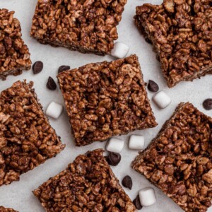Chocolate rice krispie squares on a marble table with chips and mini marshmallows scattered on the table.