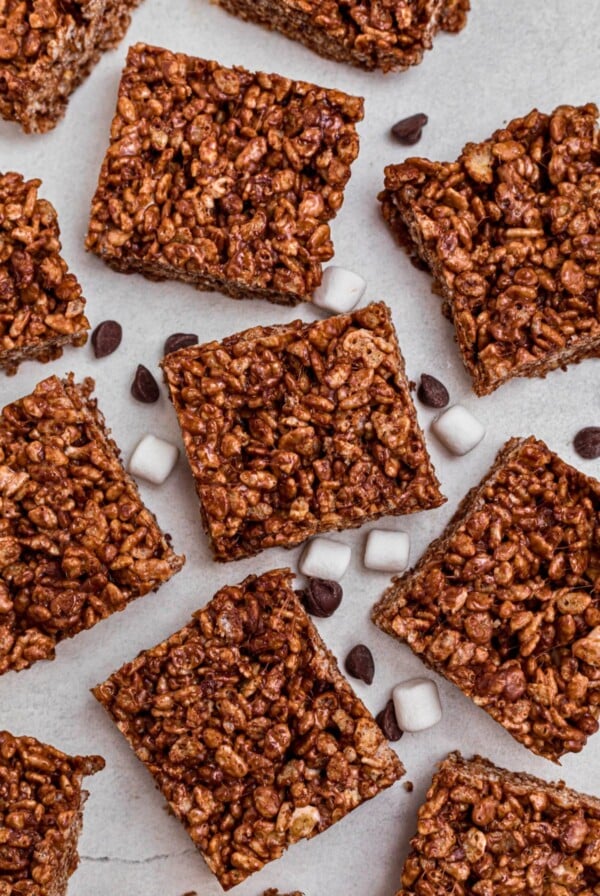 Chocolate rice krispie squares on a marble table with chips and mini marshmallows scattered on the table.