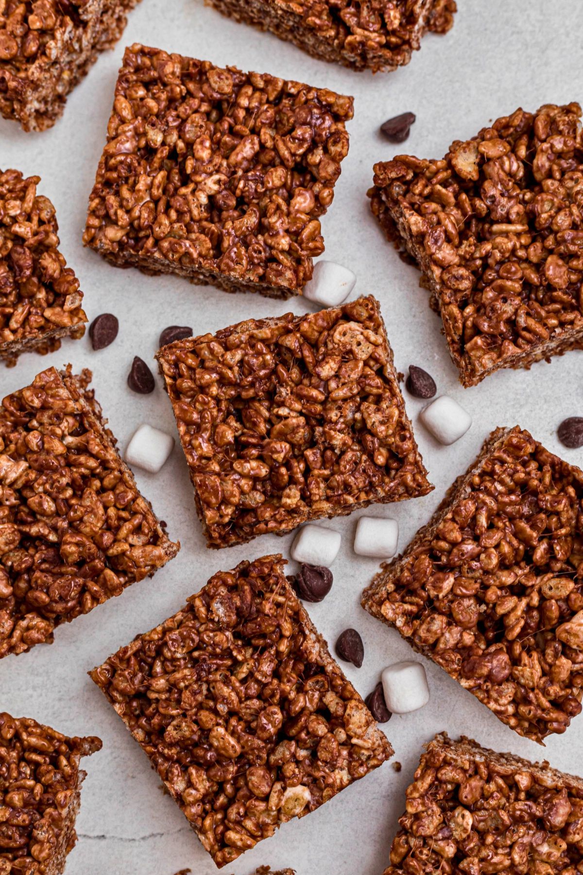 Chocolate rice krispie squares on a marble table with chips and mini marshmallows scattered on the table. 