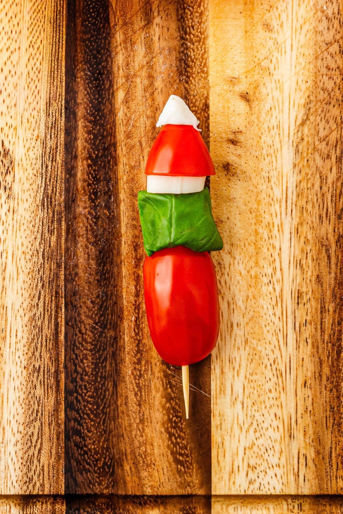Cherry tomato with basil leaf, mozzarella pieces, and string cheese pieces, on a wooden skewer, on a cutting board.