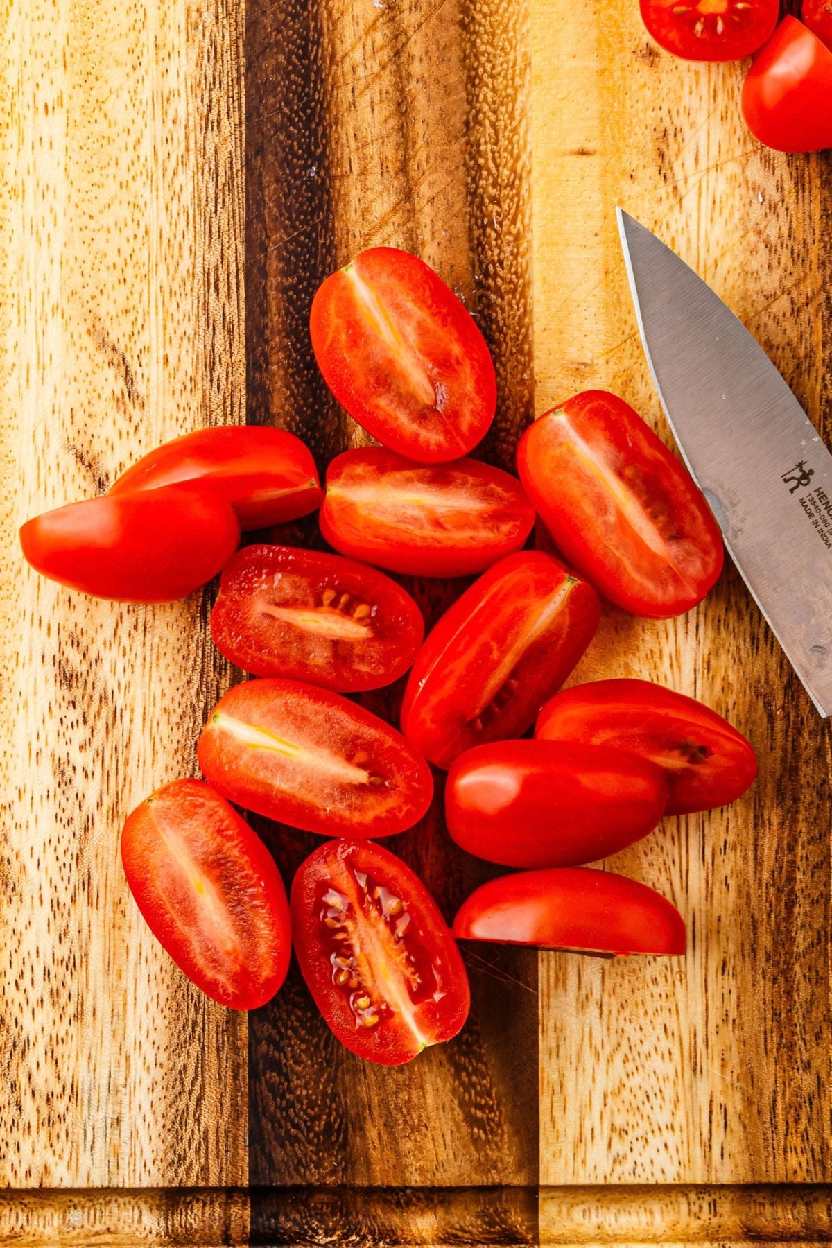 Cherry tomatoes cut in half lengthwise, on a wooden cutting board, with a knife.