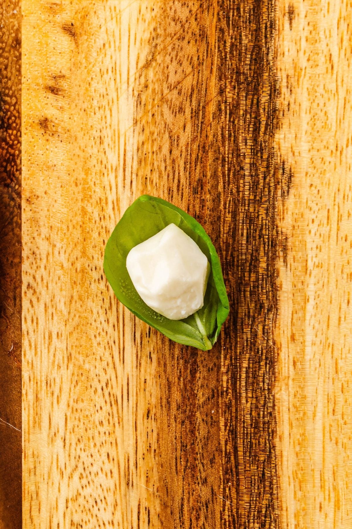 Small mozzarella pearl being wrapped basil leaf on a wooden cutting board.