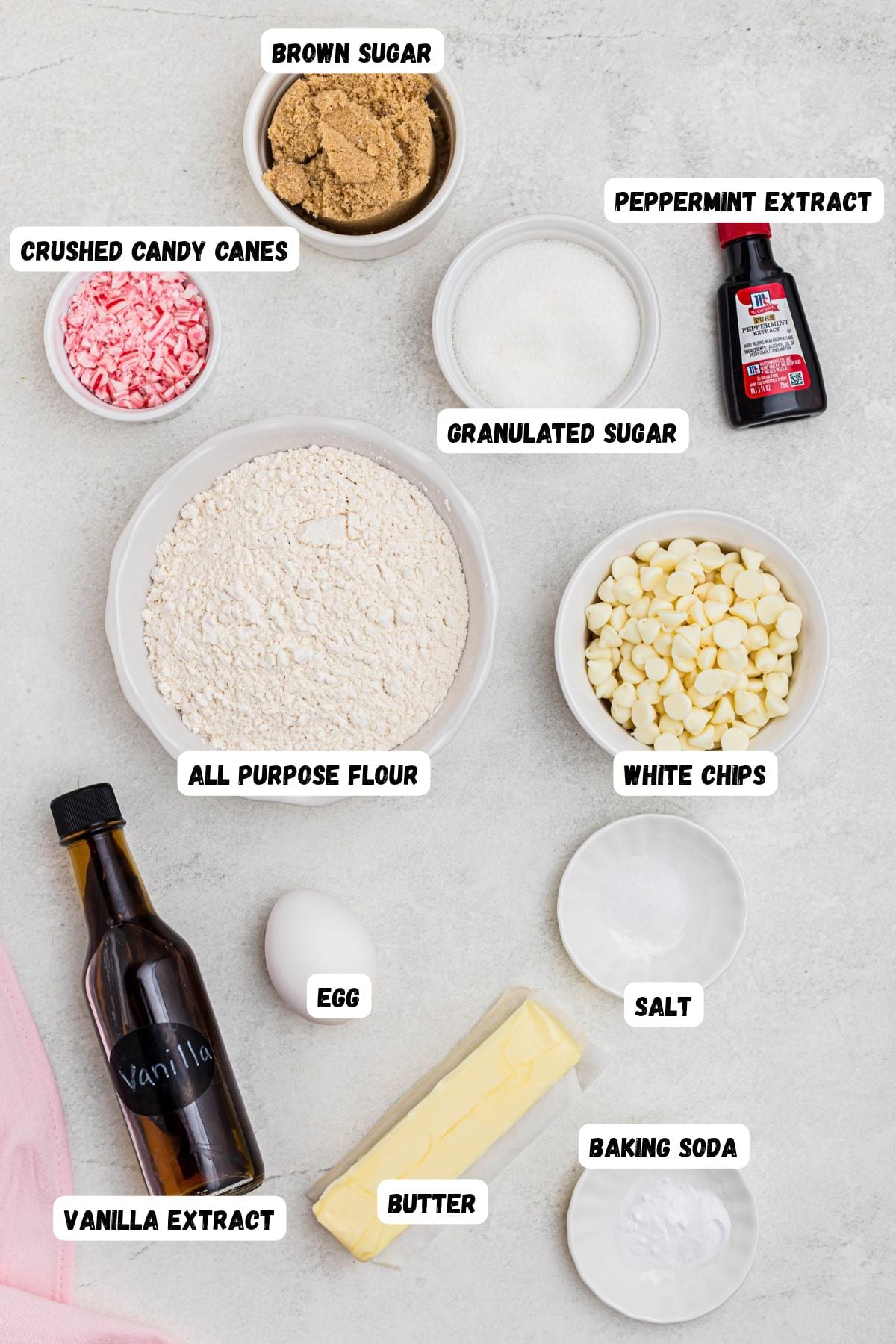 Ingredients of flour, sugars, butter, egg, extracts, butter, white chips, crushed candy cane, in white bowls, labeled, on a marble table. 